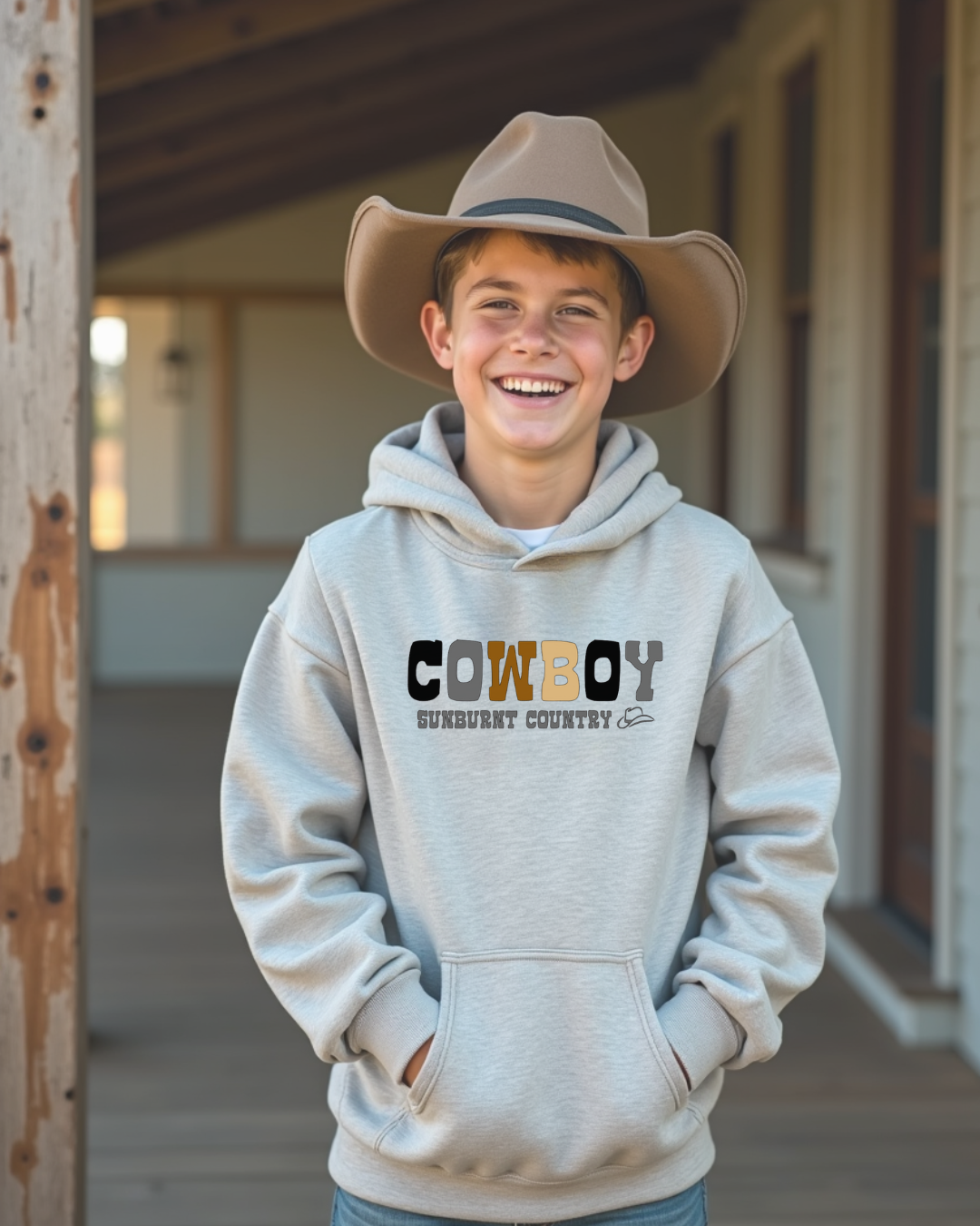 Front of a young cowboy wearing a grey coloured boys country western hoodie with the words cowboy in different coloured lettering design by Sunburnt Country Clothing, Australian kids country clothing
