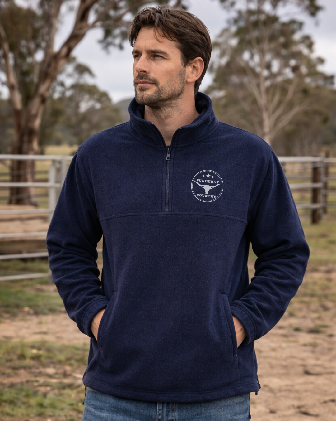 Man wearing a Sunburnt Country Clothing navy blue polar fleece jumper with a longhorn logo, standing in an outdoor setting.