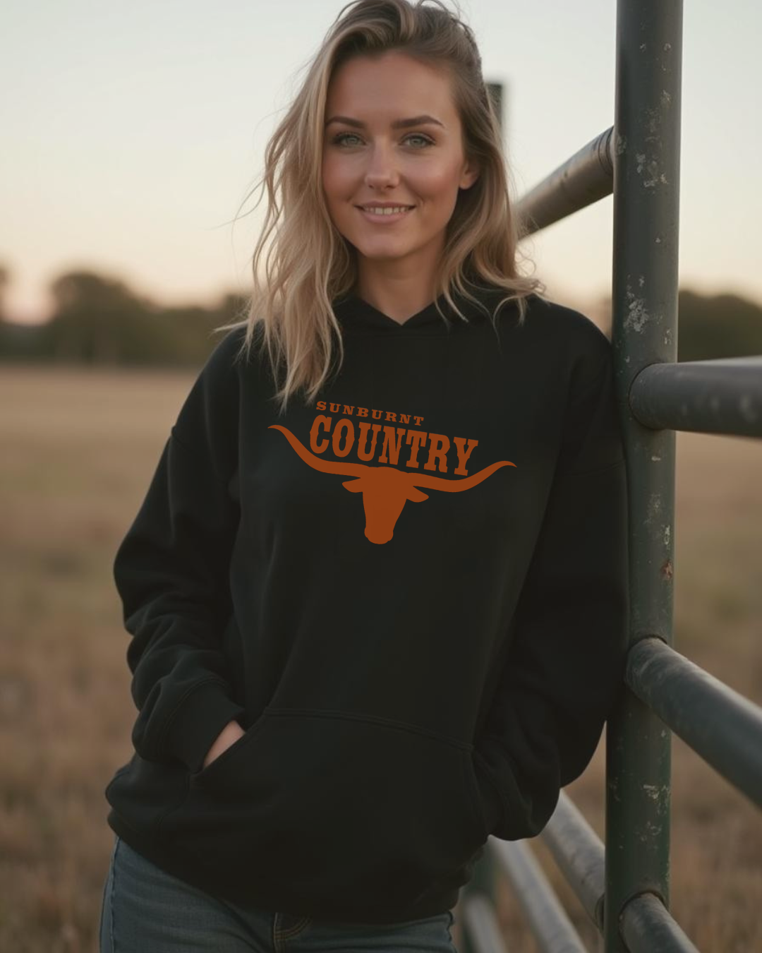 Woman wearing a black hoodie with 'Sunburnt Country' text and longhorn logo, standing in a field.