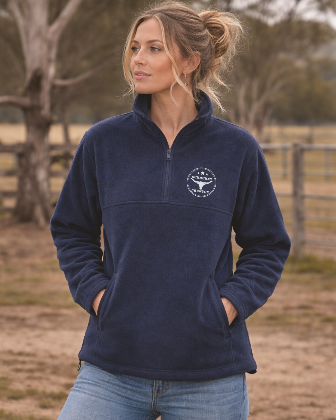 Woman wearing a Sunburnt Country Clothing navy blue polar fleece jumper with a longhorn logo, standing in an outdoor setting.