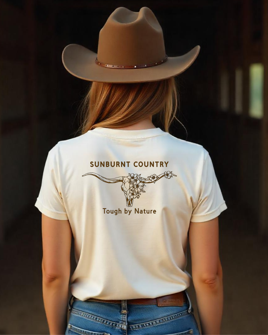 Female model wearing Sunburnt Country Clothing sand t-shirt with floral longhorn design on the back in rural setting