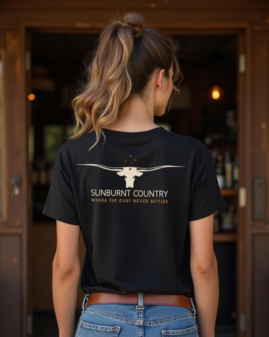 Back of Female model wearing Sunburnt Country Clothing Where the dust never settles longhorn tee in rural setting
