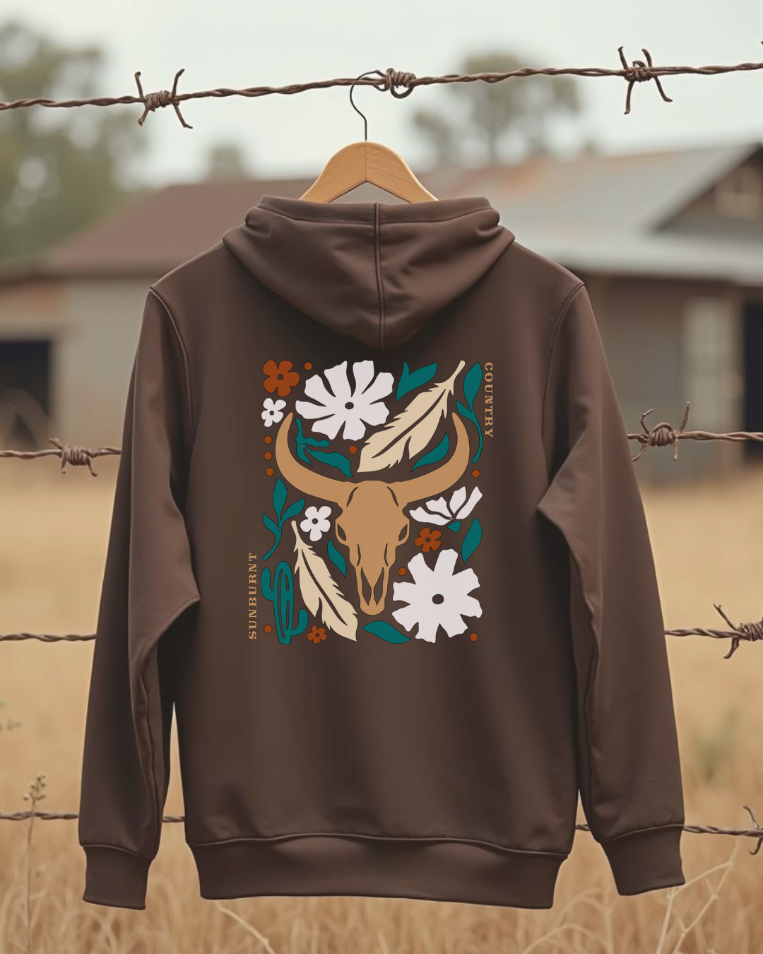 Sunburnt Country Clothing brown hoodie with flowers and longhorn skull design on the back hanging on barbed wire fence